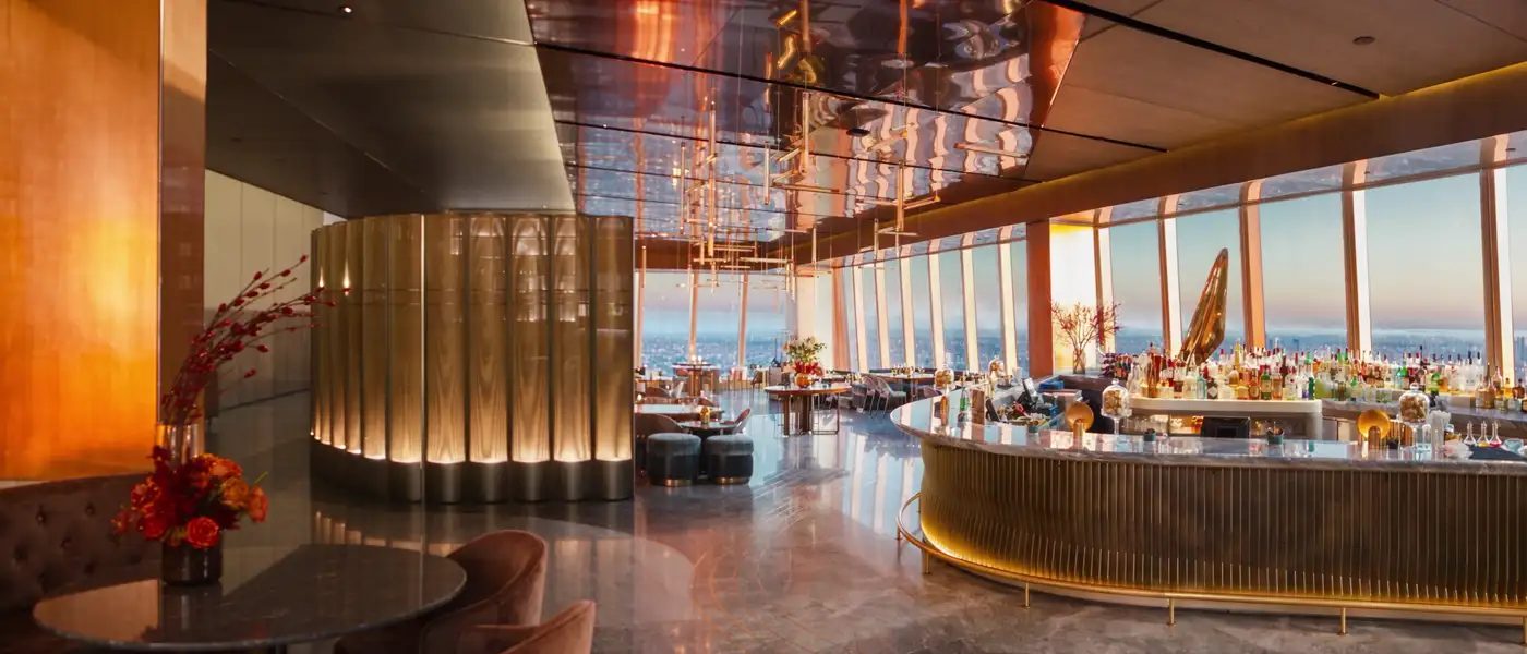 Le bar du restaurant Peak, au 30 Hudson Yards à New York : une colonne cannelée de laiton, le comptoir courbe éclairé par-dessous, la baie vitrée et Manhattan au coucher du soleil
