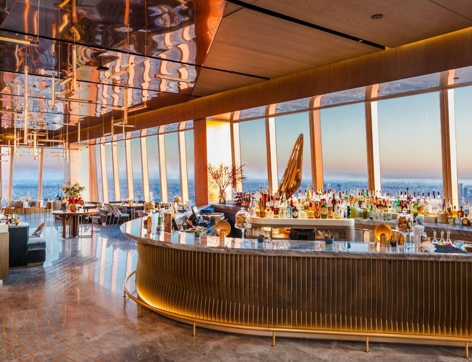 Le bar du restaurant Peak au 30 Hudson Yards : comptoir cannelé doré, plafond de bois, Manhattan au coucher du soleil derrière les vitres